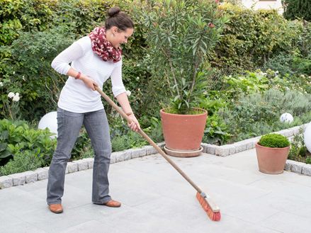 Eine Frau reinigt eine Granitterrasse mit dem Besen, im Hintergrund ein naturnaher Garten