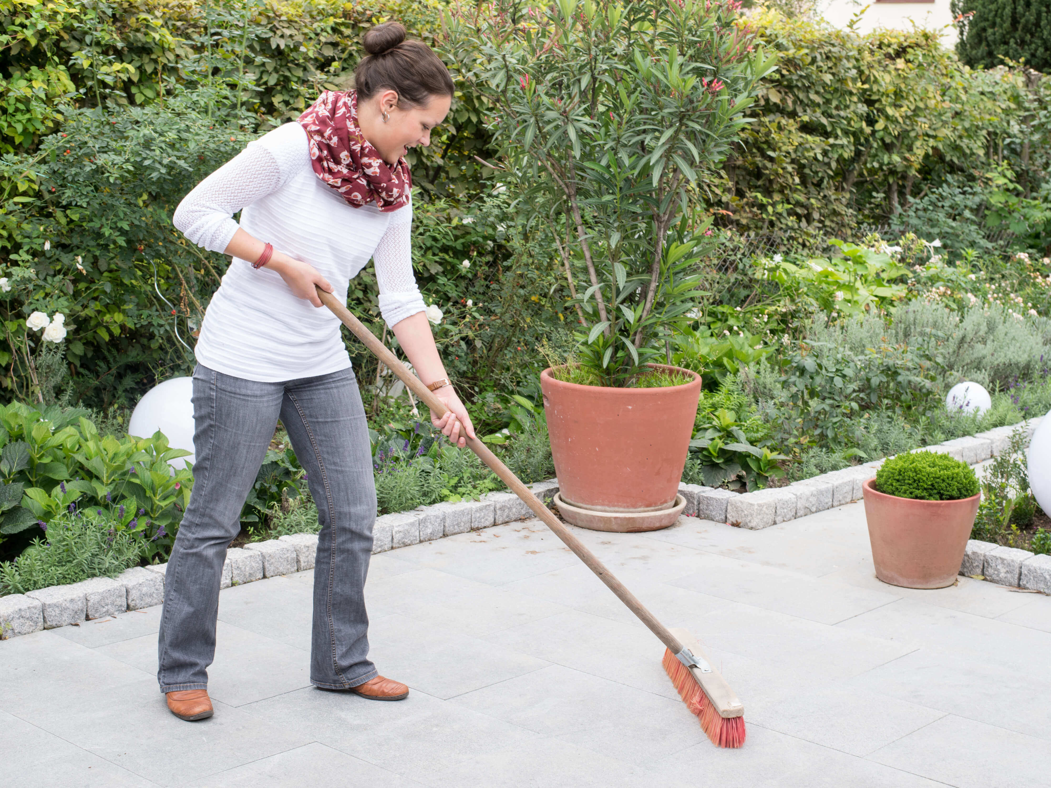 Eine Frau reinigt eine Granitterrasse mit dem Besen, im Hintergrund ein naturnaher Garten