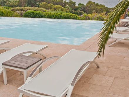 Weiße Liegestühle auf Travertinboden an einem Pool, von rechts ragt ein Palmzweig ins Bild