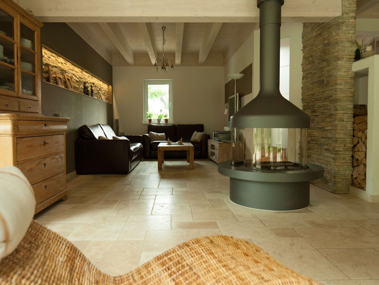 Wohzimmer mit alleinstehendem Kamin und Sitzecke auf Travertinfliese Light, im Vordergrund ein Korbsessel